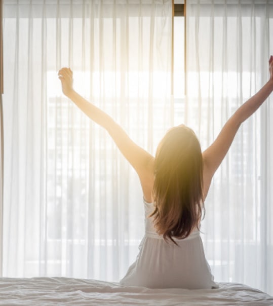 Frau in weißem Nachthemd streckt sich auf Bett während Morgensonne durch Fenster scheint
